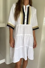 Vestido corto blanco de lino, con mangas amplias y escote en V con botones. Presenta detalles de bandas en tonos beige y negro con puntillas decorativas en el frente y en los puños. Diseño de falda con volados escalonados y silueta holgada.