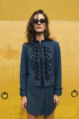 Blazer corto de tweed en color azul, con cuello mao y manga larga. Presenta detalles de pasamanería negra en el frente y cierre con ganchos metálicos y falsos botones dorados.