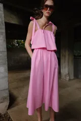 Vestido midi de color rosado con escote recto elástico y tirantes anchos con lazos ajustables. Presenta cintura elástica y bolsillos laterales ocultos en la costura.