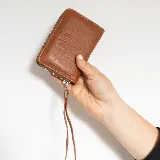 Billetera de cuero color marrón, con cierre perimetral, correa de mano incorporada y logo grabado de la marca "Del Sur" en el frente.