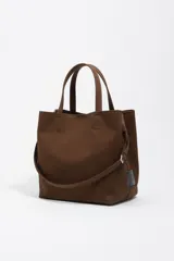 Bolso tipo tote de Bimba y Lola en color marrón, confeccionado en gabardina con detalles en piel. Cuenta con doble asa de mano, asa bandolera regulable, cierre con botón de presión y logo metálico aplicado en el frente.