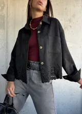 Chaqueta negra de gamuza con cuello camisero, cierre frontal de botones y detalle de bordado calado en el ruedo. Presenta mangas largas con puños abotonados y bolsillos frontales tipo parche.