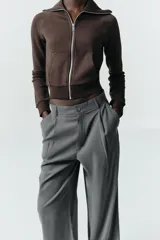 Pantalón gris de tiro alto con bolsillos delanteros y traseros, detalle de pliegues frontales y cierre frontal con cremallera y botón.