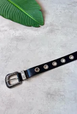 Cinturón negro de cuero sintético con hebilla plateada de estilo western ornamentada y ojales metálicos plateados a lo largo de la tira.