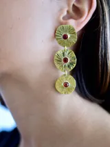 Caravanas colgantes de bronce con diseño de tres discos texturizados en degradé, cada uno con una circonia roja engarzada en el centro. Cuentan con perno de plata 925.