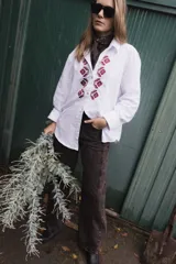 Camisa de algodón color marrón con corte clásico y cuello camisero. Presenta un bordado geométrico en color negro en la parte frontal y mangas largas con puños abotonados.