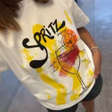 Remera blanca de corte clásico con estampa frontal ilustrada que representa un cóctel Spritz, incluyendo texto alusivo y detalles en tonos amarillos, naranjas y rojos con efecto de pinceladas.