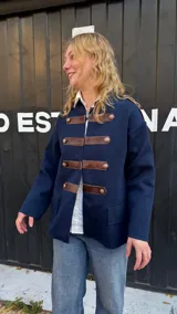 Campera de estilo militar en color azul marino, confeccionada en tejido de punto grueso. Presenta un diseño de cuello redondo, cierre frontal con tiras de cuero marrón y botones metálicos decorativos. Posee un corte recto y holgado.