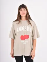 Remera de corte oversize color beige, con cuello redondo y mangas cortas. Presenta un estampado frontal con la frase 'Amore Mio', un dibujo de dos cerezas y la leyenda 'South Tuscany Italy'.