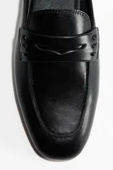 Mules estilo mocasín de cuero negro, con punta de almendra y detalle de tira penny con perforaciones decorativas en el empeine. Diseño destalonado con forro de punto.