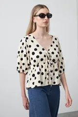 Blusa de corte relajado con estampado de lunares negros sobre fondo beige. Presenta escote en V con lazos frontales ajustables, mangas cortas abullonadas con puños elásticos y detalle de frunce bajo el busto que aporta movimiento.