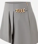 Short pollera gris de corte sastrero, confeccionado en crepe de poliéster y elastano. Presenta pliegues delanteros y un detalle de cadena metálica dorada en el lateral de la cintura.