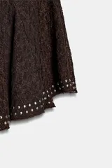 Falda corta de tiro medio confeccionada en tejido de encaje color marrón, con diseño evasé y detalle de aplicación de tachas metálicas en la cintura y el ruedo.