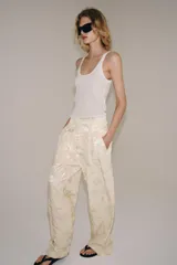Pantalón de tiro medio confeccionado en hilatura con lino, con cintura con trabillas, bolsillos laterales ocultos y pierna ancha. Presenta un diseño con estampado jacquard de motivos florales en tonos beige y crudo, y cierre frontal con cremallera, botón y ganchos metálicos.
