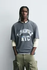Camiseta de corte boxy fit con cuello redondo y manga corta. Presenta un diseño con efecto lavado y estampado gráfico frontal con tipografía universitaria.