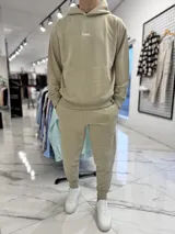 Conjunto de jogging beige que incluye un hoodie con capucha, bolsillo frontal tipo canguro y pequeño texto estampado en el pecho, acompañado de un pantalón tipo jogger con elástico en los tobillos.