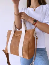 Cartera tipo tote de cuero color camel con detalles de bandas verticales en color crema. Cuenta con doble asa de mano, correa larga ajustable y extraíble, y cierre superior. Incluye etiqueta de cuero con logo grabado en el frente.