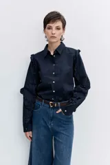 Camisa de color azul oscuro, con cuello clásico, cierre frontal de botones y detalle de volados en los hombros y a lo largo de las mangas largas.