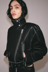 Chaqueta tipo biker de piel negra con solapas, cierre frontal asimétrico con cremallera, bolsillos con cierre y cinturón ajustable con hebilla metálica en la cintura.
