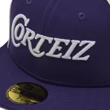 Gorra tipo cap de visera plana, colaboración entre Corteiz y New Era. Presenta un diseño bicolor en violeta y amarillo, con el logo de Corteiz bordado en blanco en el frente y el logo de New Era en el lateral.