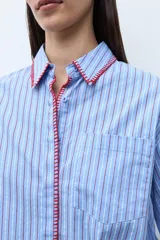 Camisa de corte clásico con estampado de rayas verticales en tonos celestes y blancos. Presenta un detalle de bordado decorativo en color rojo a lo largo de la botonadura frontal y el cuello, mangas largas con puños abotonados y un bolsillo frontal en el pecho.