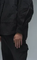 Chaqueta tipo overshirt de microfibra, color negro, con calce oversize y ruedo curvo. Presenta cierre frontal con broches a presión, bolsillos frontales de gran tamaño y cierres laterales a lo largo de las mangas para ajustar su amplitud.