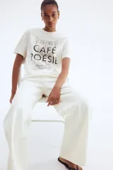 Remera blanca de manga corta y cuello redondo, con estampado gráfico en negro que simula la fachada de un café parisino con el texto "CAFÉ POÉSIE" y la dirección "Rue La Fayette, 3 Paris, France".