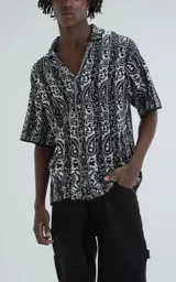 Camisa de manga corta con tejido de punto abierto tipo crochet, corte oversize y cuello solapa. Presenta un diseño estampado con rayas verticales y motivos paisley en blanco y negro.
