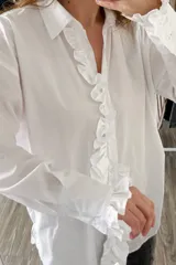 Camisa blanca de algodón con cuello clásico, cierre frontal con botones, mangas largas con puños abotonados y volados en el frente y los puños.