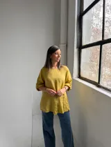 Blusa mostaza de lino, con cuello redondo, mangas tres cuartos con presilla y botón, y detalle de pliegues en la parte inferior.