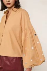 Camisa color beige de corte crop, con cuello camisero, mangas largas abullonadas con detalle de espejos y cierre frontal con botones.