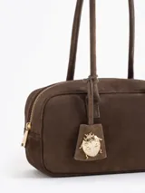 Cartera de mano de estructura rectangular, confeccionada en cuero descarne vacuno color verde oscuro. Presenta doble manija larga, cierre superior y un charm metálico dorado en forma de vaquita de San Antonio colgando de un tag lateral. Incluye el logo de la marca grabado en relieve en el frente.