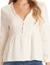 Blusa de algodón color beige con escote en V, cierre frontal con botones y detalles de puntilla en el frente. Presenta mangas largas amplias con puños elastizados y un diseño con bordados sutiles.