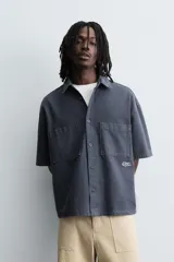 Camisa de algodón con estructura canvas, color rojo lavado, de corte relajado (relaxed fit) y manga corta. Posee cuello solapa, dos bolsillos de plastrón en el pecho y cierre frontal de botonadura. Presenta bordado y estampado de texto a contraste en el delantero.