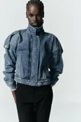 Cazadora de denim estilo crop con cuello solapa, manga larga y cierre frontal con cremallera y botón metálico. Presenta bolsillos de vivo en el delantero y detalle de trabillas en los hombros.