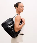 Bolsa tipo shopper de tamaño grande, confeccionada en poliuretano texturizado con acabado brillante. Presenta un diseño estructurado con correas fijas para el hombro, cierre superior mediante botón a presión y un detalle metálico decorativo en la parte frontal.