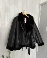 Campera tipo gamulán de cuero sintético negro con interior y detalles de corderito al tono. Presenta cierre diagonal, cuello amplio, bolsillos laterales con cierre y cinturón ajustable en la cintura.