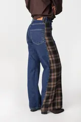 Jean de pierna ancha con diseño bicolor, combinando denim azul clásico con un panel lateral de tela a cuadros en tonos marrón y negro. Presenta cuatro bolsillos funcionales y cierre frontal.