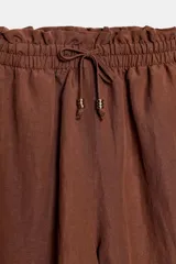 Bermuda marrón de lino, de corte holgado, con cintura elástica fruncida y cordón ajustable con abalorios en los extremos. Cuenta con bolsillos delanteros.