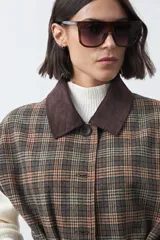 Chaleco de corte relajado con estampado a cuadros en tonos tierra, cuello camisero oscuro y cierre frontal con botones. Incluye un cinturón de la misma tela para ajustar a la cintura y bolsillos frontales.