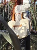 Bolso tipo tote color crema, con detalles en cuero color marrón. Tiene un bolsillo con cierre en la parte superior y un parche de cuero con el logo de la marca en la parte inferior.