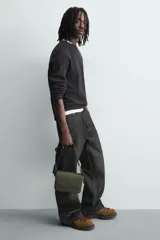 Bandolera verde militar con acabado engomado y textura geométrica. Tiene un compartimento principal con cierre de solapa y botón a presión, un bolsillo interior con cremallera y un asa de hombro ajustable con monedero.