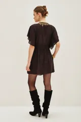 Vestido corto de color negro con escote en V profundo, adornado con bordados dorados y pedrería. Presenta mangas cortas tipo mariposa, cintura ajustable con cordón y bolsillos laterales.