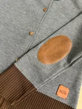 Cardigan de lana merino color gris con detalles en marrón, cuello redondo, cierre frontal con botones y coderas marrones.