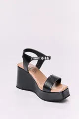 Sandalias de cuero negras con plataforma y tacón de 10 cm. Tienen una tira ancha en la parte delantera y una tira ajustable con hebilla alrededor del tobillo, ambas con detalles de tachas plateadas.
