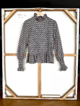 Blusa de algodón con diseño de nido de abeja en el torso, cuello alto con volado y mangas largas con puños elásticos terminados en volado. Presenta un estampado floral pequeño en tonos marrón y crudo.