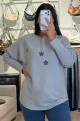 Buzo tejido de punto en color beige, con diseño de flores aplicadas en tono gris oscuro y detalles de brillos plateados que irradian desde las flores. Presenta cuello redondo amplio, mangas largas con caída holgada y corte recto.