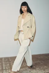 Trench corta de color beige con cuello de solapa y manga larga. Cuenta con bolsillos delanteros, cintura ajustable con cordones, bajo abullonado y cierre frontal cruzado con un botón visible. El interior está forrado con tela a cuadros.