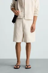 Bermuda de corte relajado color beige claro, confeccionada en tejido con textura. Presenta cierre frontal con cremallera y botón, bolsillos delanteros y bolsillos traseros de doble vivo con botón.