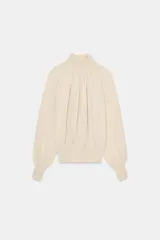 Blusa fluida de punto combinado color beige, con cuello alto, detalles de botonadura lateral en el cuello, mangas largas abullonadas y puños ajustados.